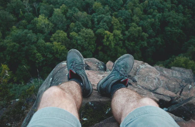 a man sitting on top of a rock next to a forest, trending on pexels, detailed shot legs-up, sneaker photo, as seen from the canopy, standing on the mast