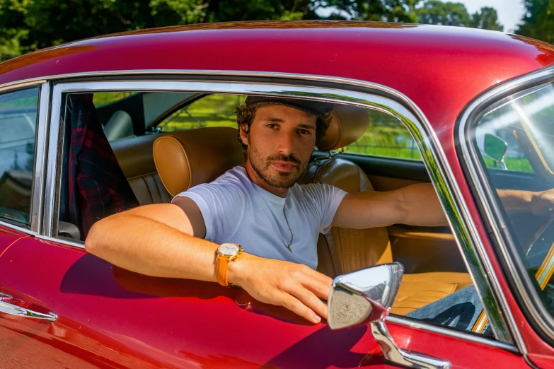 a man sitting in the driver's seat of a red car, a portrait, bulgari style, daniel ricciardo, dressed in 1970s menswear, stained antique copper car paint