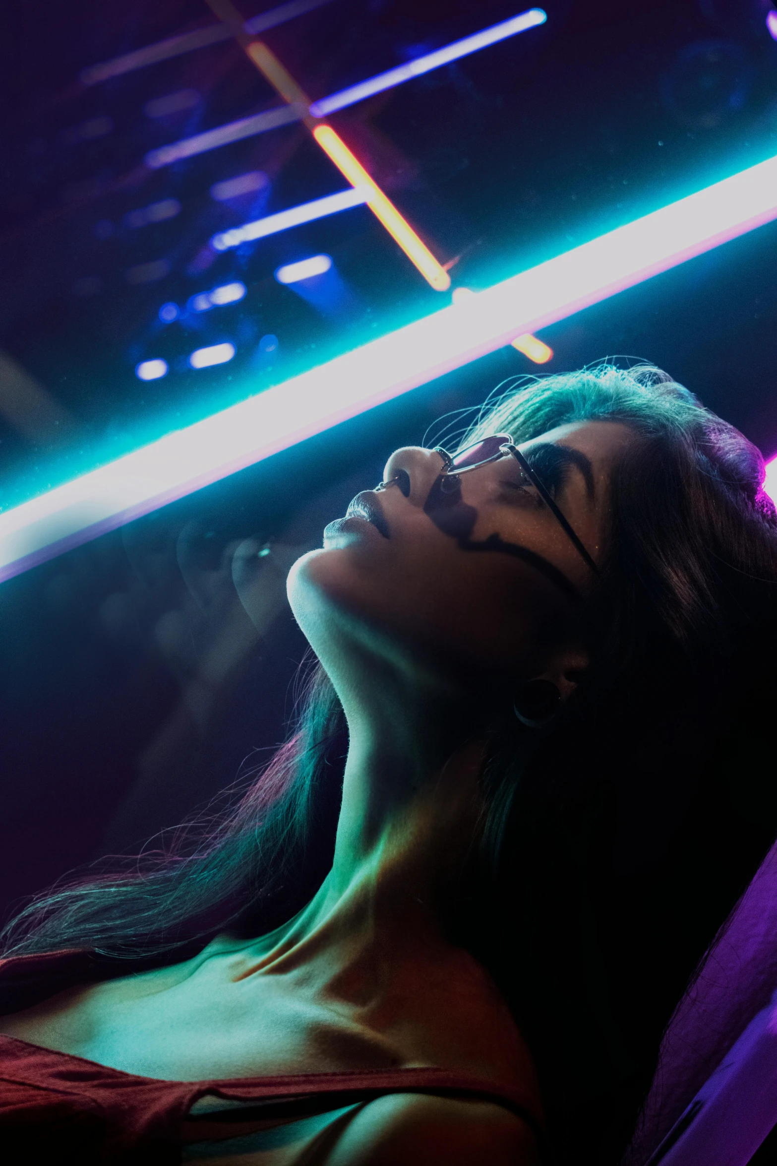 a woman sitting on a couch in front of a neon light, inspired by Elsa Bleda, trending on pexels, holography, in a strip club, girl with glasses, as she looks up at the ceiling, backlit beautiful face