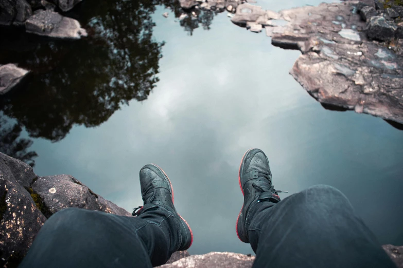 a person standing on top of a rock next to a body of water, by Alexander Brook, pexels contest winner, realism, wearing black boots, view from below, sitting at a pond, sneaker photo