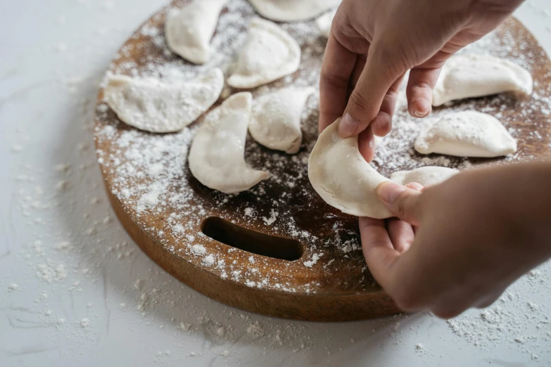 a wooden cutting board topped with dumplings covered in powdered sugar, inspired by Géza Dósa, mingei, hands, thumbnail, half moon, handcrafted