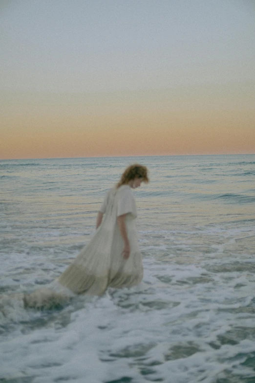 a woman in a white dress standing in the ocean, an album cover, unsplash, renaissance, back of emma stone in beige coat, pale skin curly blond hair, at dusk at golden hour, ignant