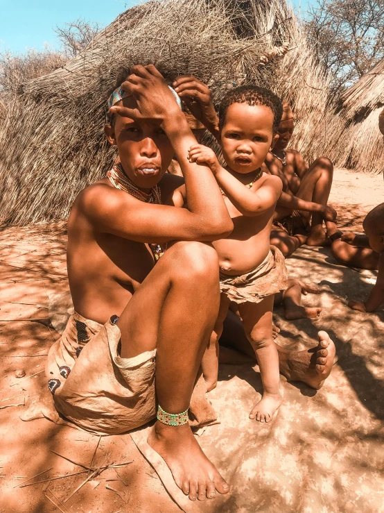 a group of people sitting on top of a dirt field, by Hubert van Ravesteyn, pexels contest winner, afrofuturism, facial tribal markings, father with child, wearing cave man clothes, instagram story