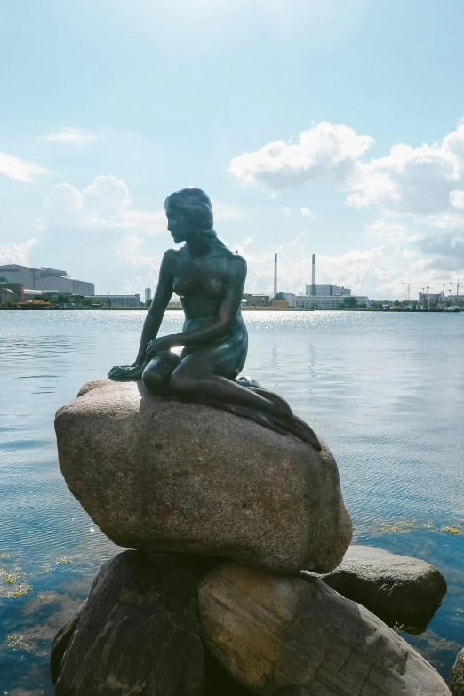 a statue of a mermaid sitting on top of a rock, inspired by Wilhelm Marstrand, at the waterside, in the distance, iconic scene, made of rubber