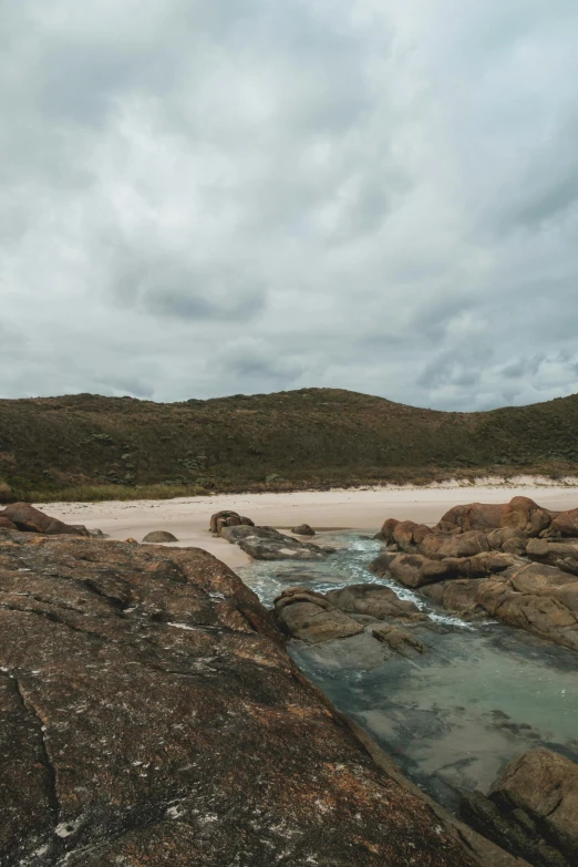 a man standing on top of a rock next to a body of water, australian beach, gloomy skies, dry river bed, white sand beach