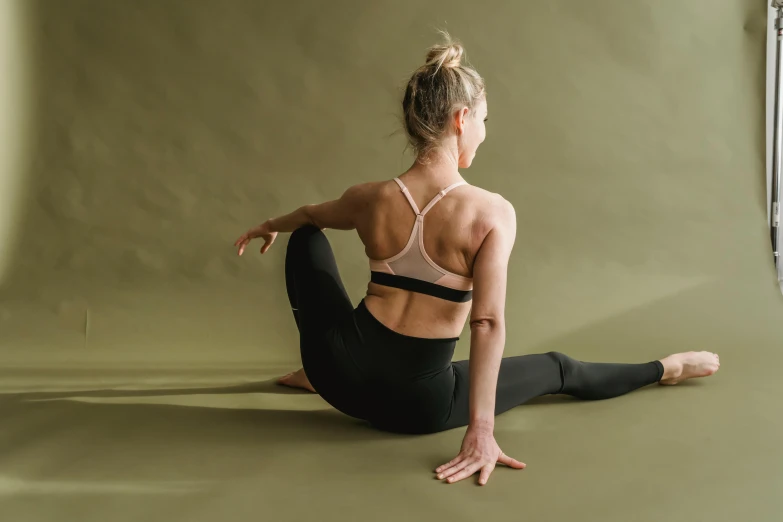 a woman sitting on the ground in a yoga pose, trending on pexels, arabesque, showing her shoulder from back, tights skin, looking towards camera, manuka
