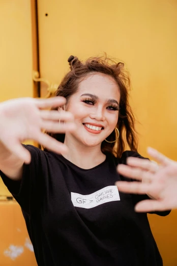 a woman standing in front of a yellow wall, a picture, trending on pexels, wearing a black shirt, hand gesture, south east asian with round face, concert photo