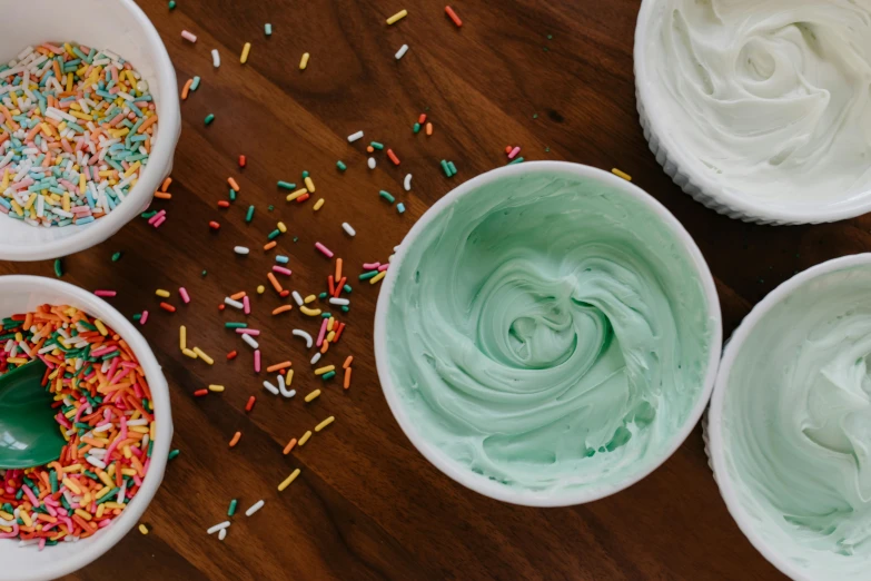 a wooden table topped with bowls of frosting and sprinkles, inspired by Wayne Thiebaud, trending on pexels, sea foam, geeen skin, yogurt, green