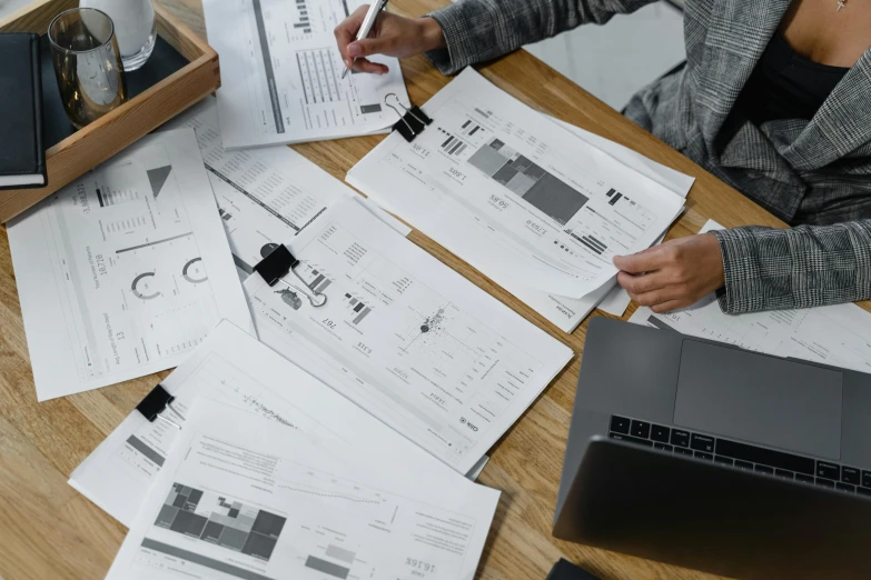 a woman sitting at a table with papers and a laptop, a cartoon, pexels contest winner, engineering blueprints, informative graphs and diagrams, a high angle shot, selling insurance