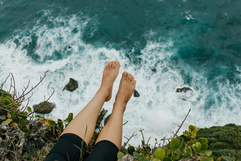 a person sitting on top of a cliff next to the ocean, barefeet, natural skin, taking from above, weightless