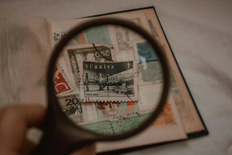 a person holding a magnifying glass over a book, a microscopic photo, by Emma Andijewska, pexels contest winner, vintage 1950s stamp, crafts and souvenirs, viewed from bird's-eye, postage