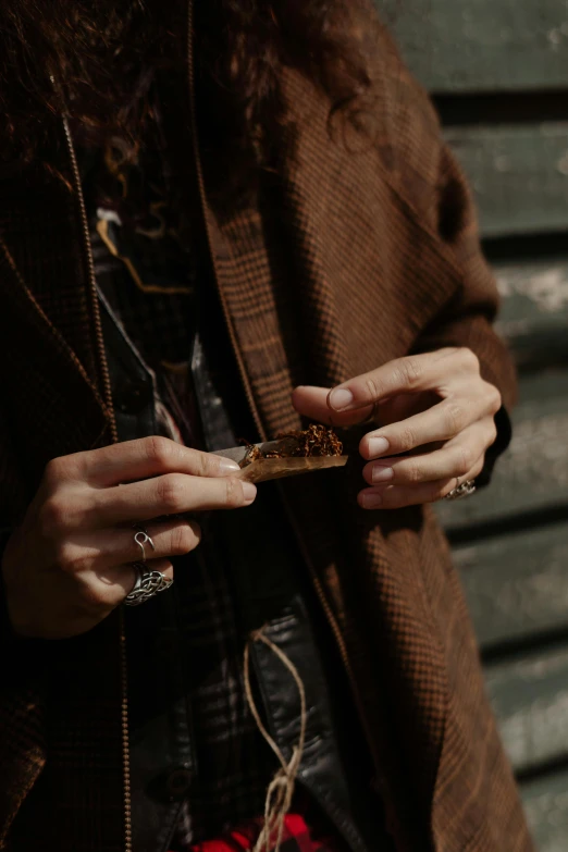 a close up of a person holding a cell phone, holding a cigar, dried plants, brown jacket, hippie