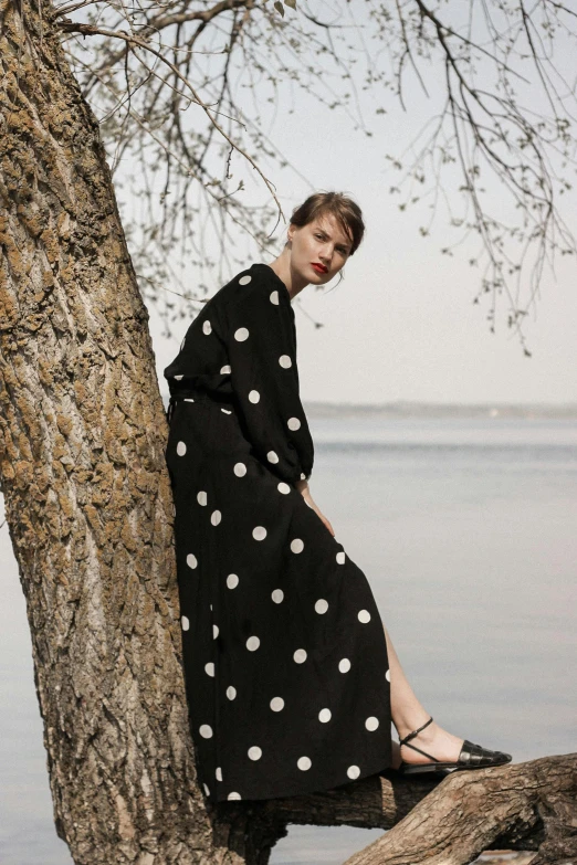 a woman is sitting on a tree by the water, a portrait, unsplash, polka dot, sleek robes, comme des garcon campaign, tall shot