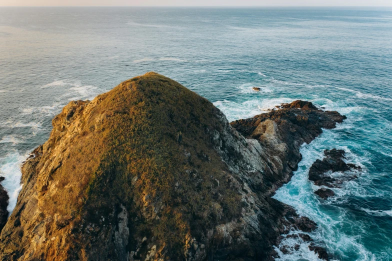 a large rock in the middle of a body of water, hills and ocean, helicopter view, profile image, golden hour photo