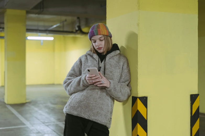 a woman standing in a parking garage looking at her cell phone, inspired by Elsa Bleda, trending on pexels, graffiti, knitted hat, beige hoodie, julia garner, yellow carpeted