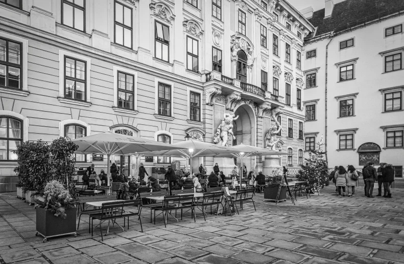 a black and white photo of a courtyard with tables and umbrellas, a black and white photo, by Emma Andijewska, city of munich!!!, people watching, baroque detailed, cafe