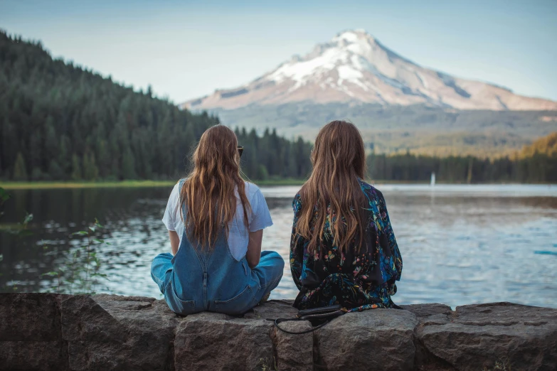 two women sitting on a rock overlooking a lake with a mountain in the background, trending on pexels, long fine hair, teenager hangout spot, on a canva, overlooking