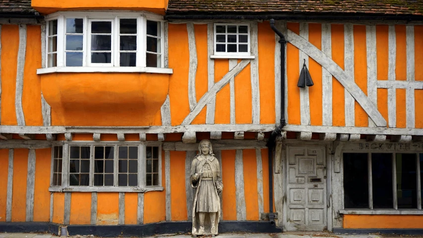 a statue of a man standing in front of a building, inspired by Edmund Greacen, pexels contest winner, arts and crafts movement, orange theme, medieval tumbledown houses, elstree, press shot