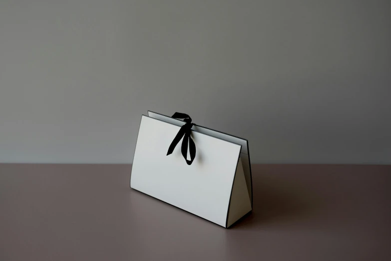 a white bag sitting on top of a table, by Eden Box, minimalism, ribbon, sleek black and white, facing front, drawfolio