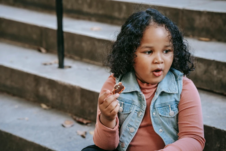 a little girl sitting on the steps eating a donut, by Nina Hamnett, pexels contest winner, she has olive brown skin, chocolate. rugged, brown curly hair, surprised expression on her face