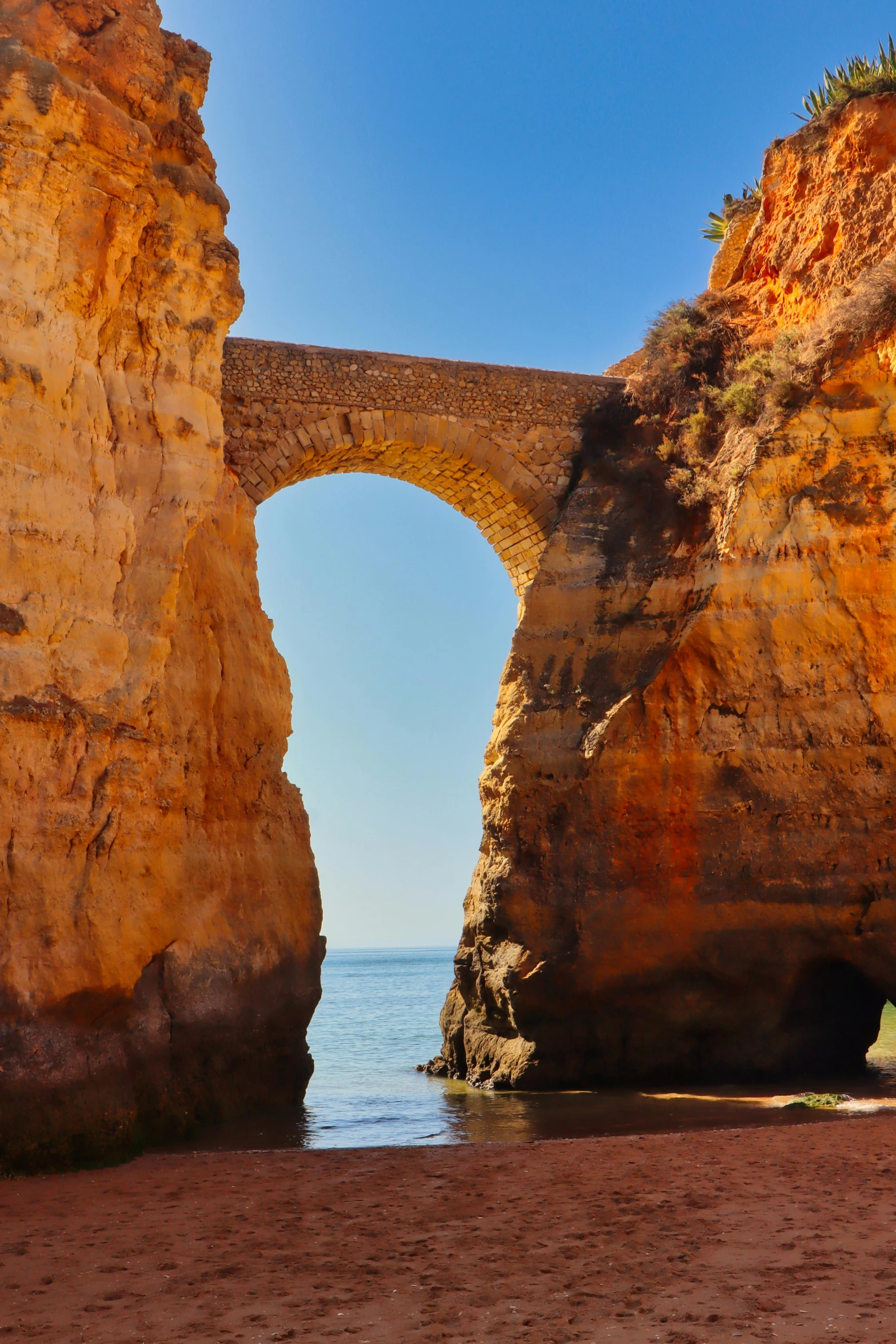 a group of people standing on top of a sandy beach, rock arches, bridge over the water, rocha, up-close