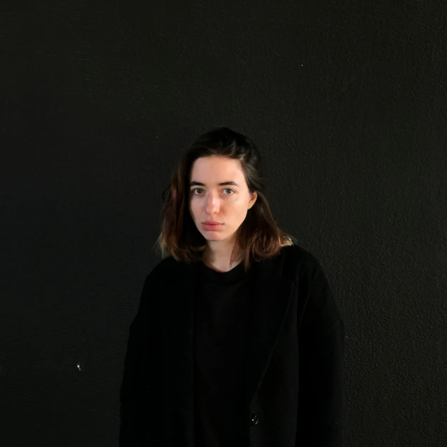 a woman standing in front of a black wall, wearing a black sweater, mateo dineen, on a pale background, portrait n - 9