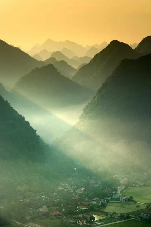 an aerial view of a valley with mountains in the background, inspired by Pierre Pellegrini, sunshafts, vietnam, light green mist, light show