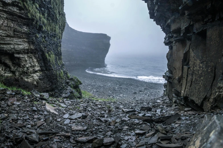 a cave filled with lots of rocks next to a body of water, by Þórarinn B. Þorláksson, pexels contest winner, les nabis, orkney islands, under a gray foggy sky, beach is between the two valleys, layers of strata
