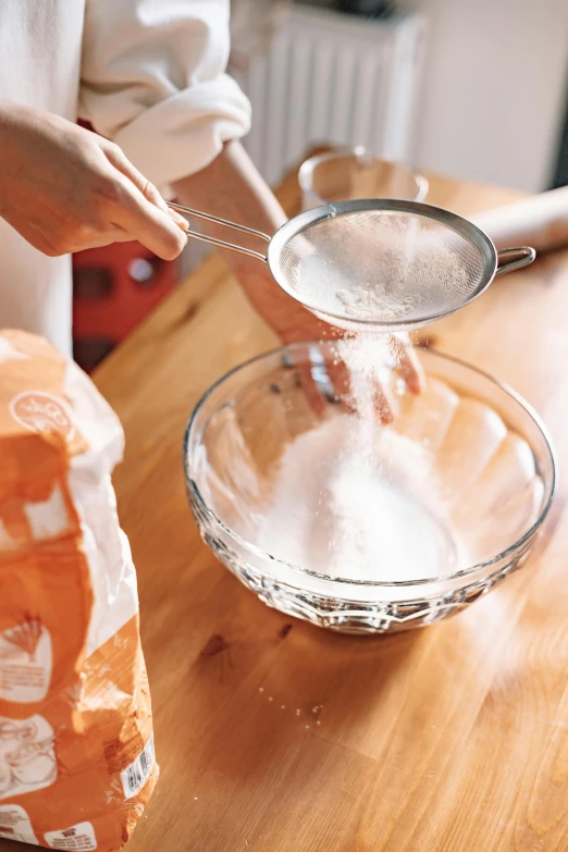 a person pouring flour into a bowl on top of a wooden table, glassware, thumbnail, plastic wrap, epicurious