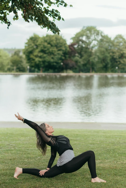 a woman doing a yoga pose in front of a lake, by Tom Bonson, arabesque, in the park, next to a tree, lena oxton, meadows