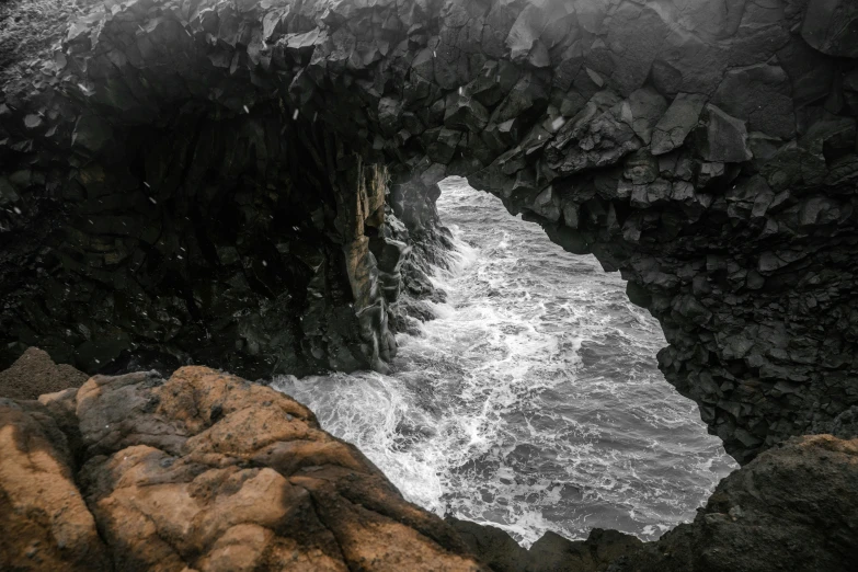 a man standing on top of a rock next to a body of water, a picture, by Matija Jama, pexels contest winner, vorticism, stone gate to the dark cave, inside the curl of a wave, black sand, barnacle