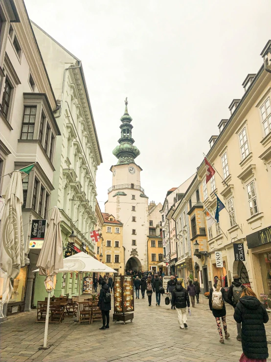 a group of people walking down a street next to tall buildings, art nouveau, neoclassical tower with dome, austrian architecture, green alleys, 💣 💥💣 💥