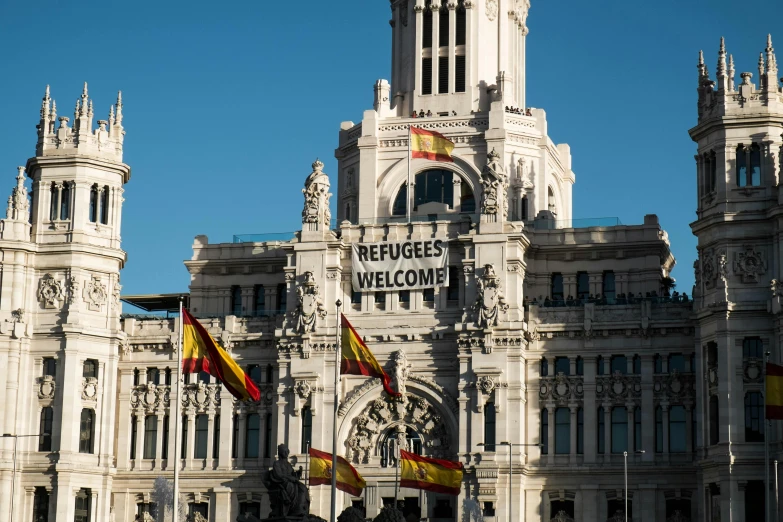 a large white building with a clock tower, by Luis Molinari, red and black flags waving, red yellow flag, refugees, thumbnail