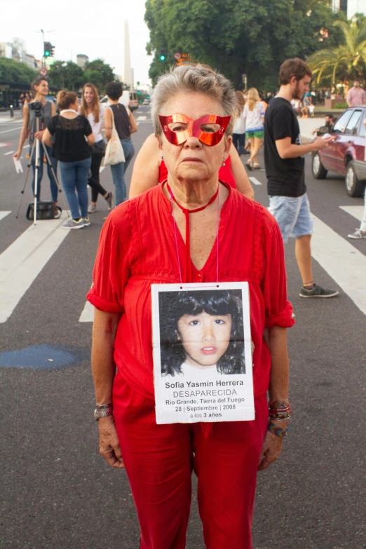 a woman standing in the middle of a street holding a sign, by Glòria Muñoz, dada, wearing red tainted glasses, older woman, mugshot, selena quintanilla perez