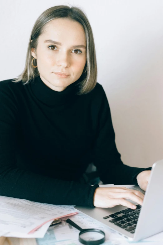 a woman sitting at a desk using a laptop computer, a portrait, inspired by Louisa Matthíasdóttir, trending on unsplash, wearing a black sweater, looking directly at the camera, danila tkachenko, professional profile picture