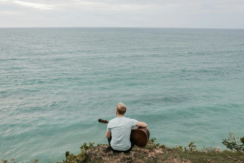 a man sitting on top of a cliff next to the ocean, an album cover, by Jessie Algie, pexels contest winner, happening, acoustic guitar, looking off to the side, turquoise ocean, lachlan bailey