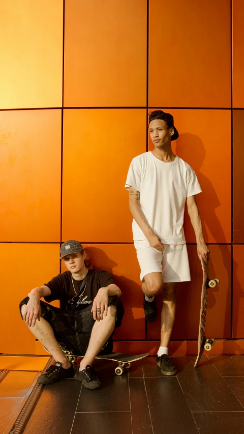 a man sitting on top of a skateboard next to a woman, by Andrew Stevovich, in front of an orange background, lee griggs and jason chan, wearing shorts and t shirt, profile image