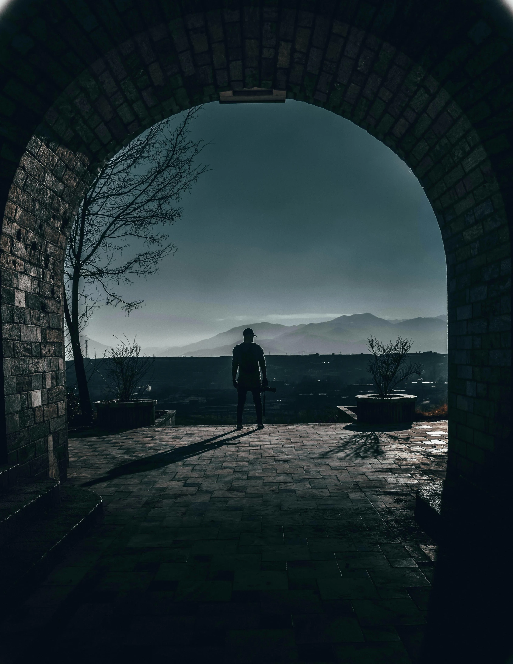 a person standing at the end of a tunnel, by Kristian Kreković, castle in the distance, with mountains in the distance, cinematic outdoor lighting, distant full body view