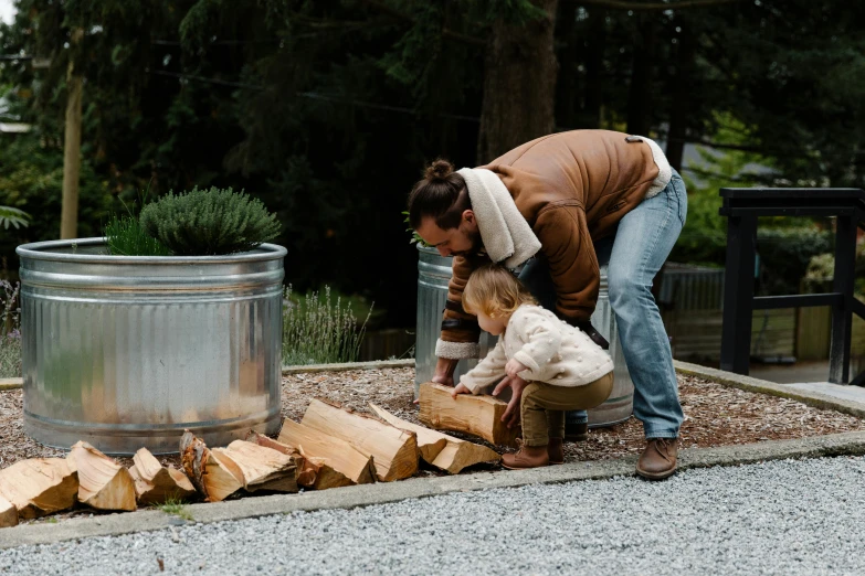 a woman standing next to a child near a pile of wood, by Jessie Algie, pexels contest winner, fire pit, denim, dwell, feed troughs