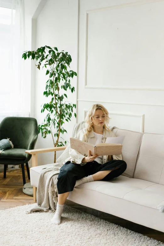 a woman sitting on a couch reading a book, inspired by Constantin Hansen, pexels contest winner, inspect in inventory image, in white room, large potted plant, a blond