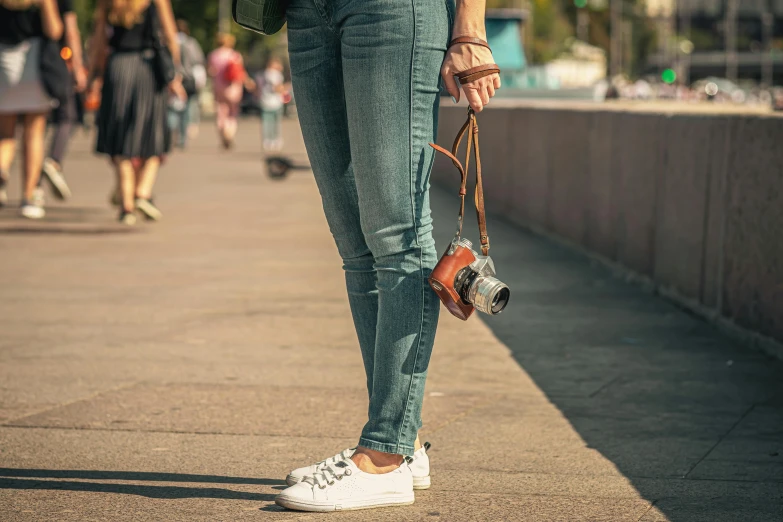 a woman standing on a sidewalk holding a camera, pexels contest winner, ( ( ( wearing jeans ) ) ), fan favorite, afternoon, sneaker photo