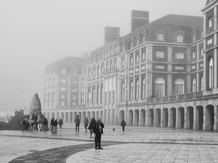 a black and white photo of people walking down a street, a black and white photo, inspired by Henri Cartier-Bresson, tonalism, standing next to royal castle!!!, ground covered in mist, photographic print, seaside