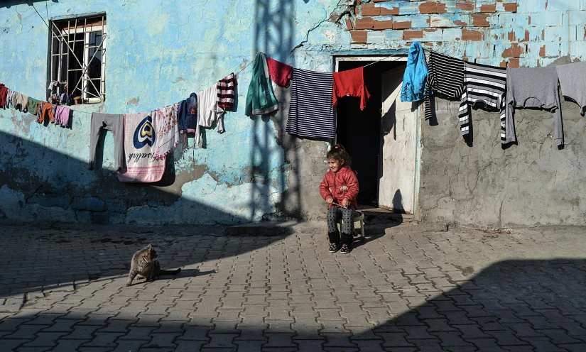 a man sitting in front of a blue building next to a cat, by Lucia Peka, pexels contest winner, kids playing, panoramic view of girl, turkey, :: morning