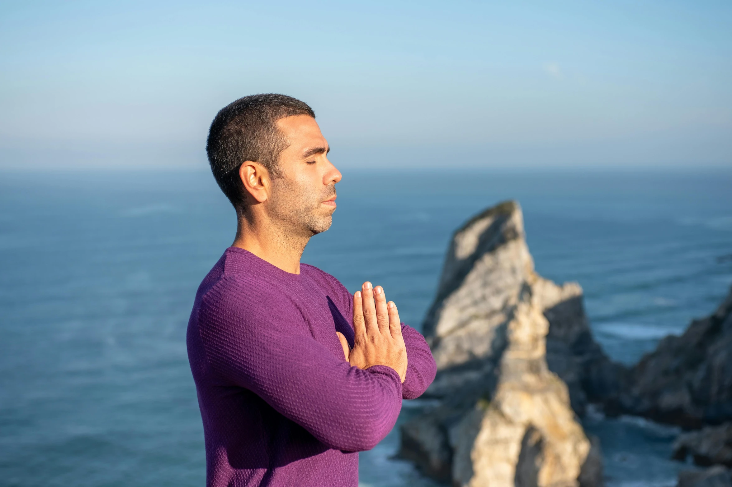 a man standing on top of a cliff next to the ocean, anjali mudra, young man in a purple hoodie, avatar image