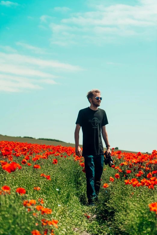 a man walking through a field of red flowers, by Simon Marmion, unsplash, color field, blonde british man, pewdiepie, near the sea, wearing orange sunglasses