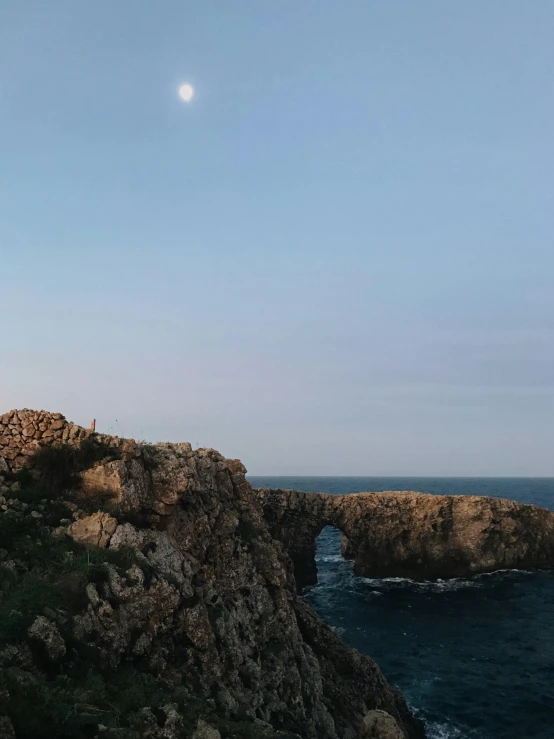 a lighthouse sitting on top of a cliff next to the ocean, crescent moon in background, panorama shot, big arches in the back, slightly minimal