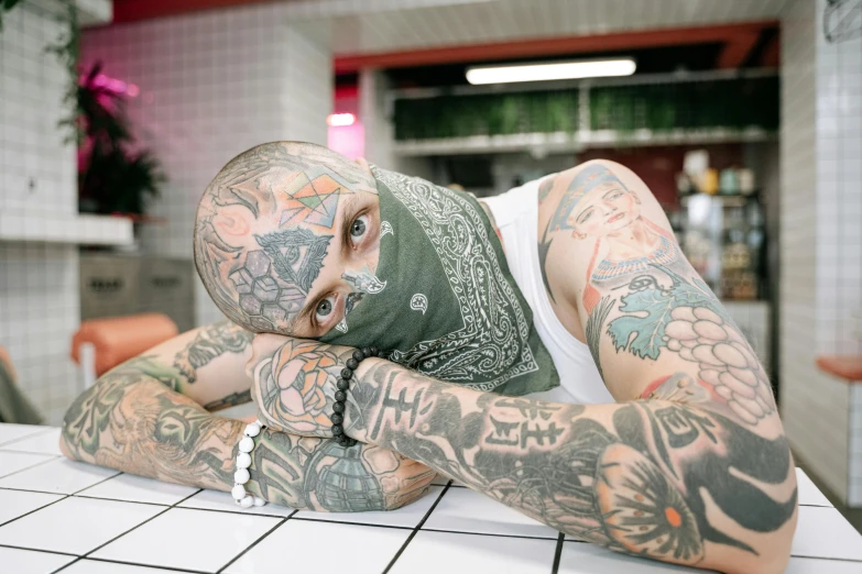 a man laying on top of a counter covered in tattoos, trending on pexels, hyperrealism, bandana, green head, food head, arms covered in gang tattoo