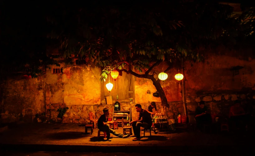 a group of people sitting around a table under a tree, by Han Gan, pexels contest winner, happening, street lanterns, musicians, vietnam, shady dark backalley at night