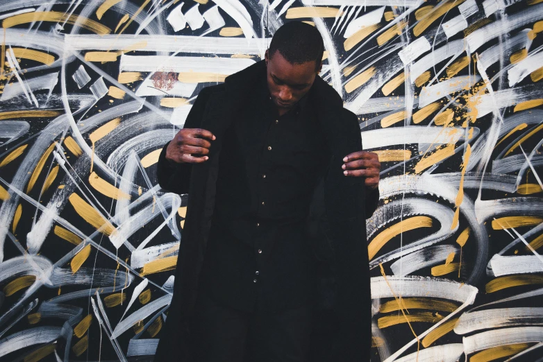 a man standing in front of a wall covered in graffiti, an album cover, inspired by Theo Constanté, lyco art, wearing black overcoat, liquid gold, ( ( dark skin ) ), intrincate clothing