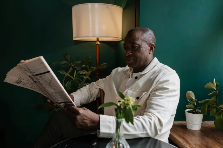 a man sitting at a table reading a newspaper, black man, gilleard james, in a dark green polo shirt, sitting in a lounge